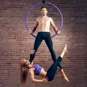X-Pole purple Aerial Hoop in front of brick wall with male model on it at whose legs a female model hangs